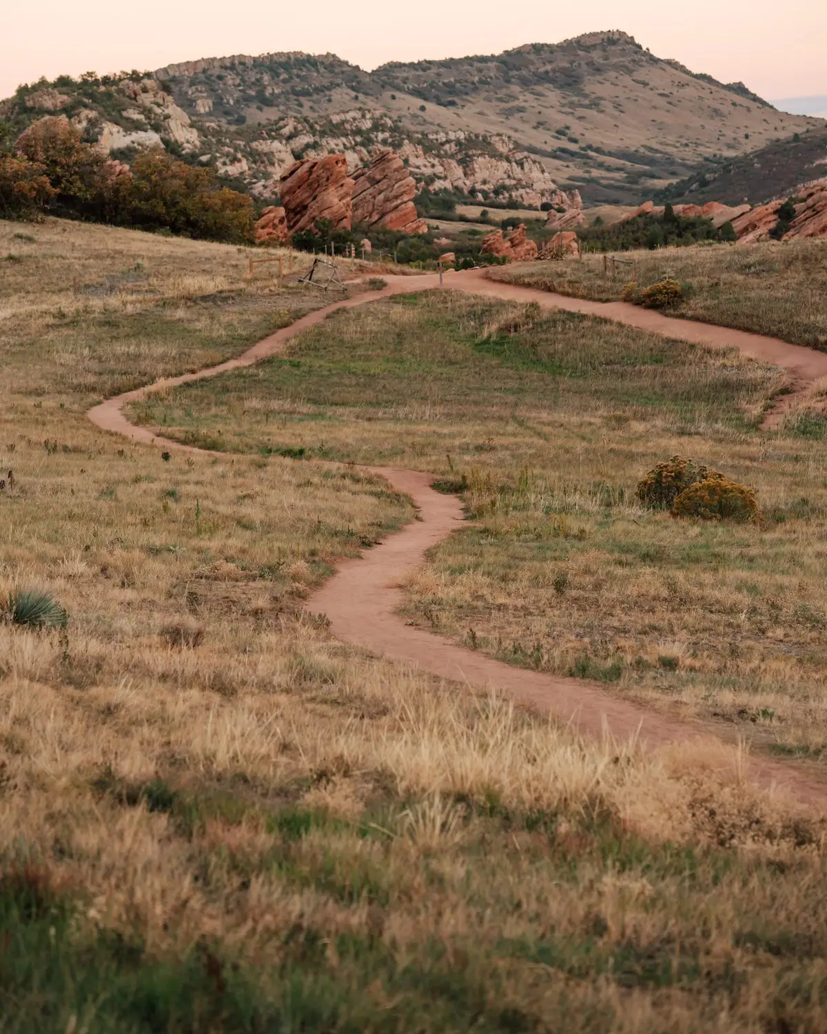 A dirt trail winding through golden grass toward red rocks and the Colorado foothills
