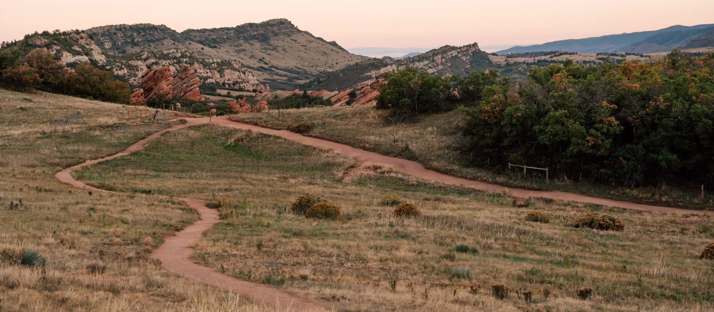 A dirt trail winding across a foothills meadow toward red rocks
